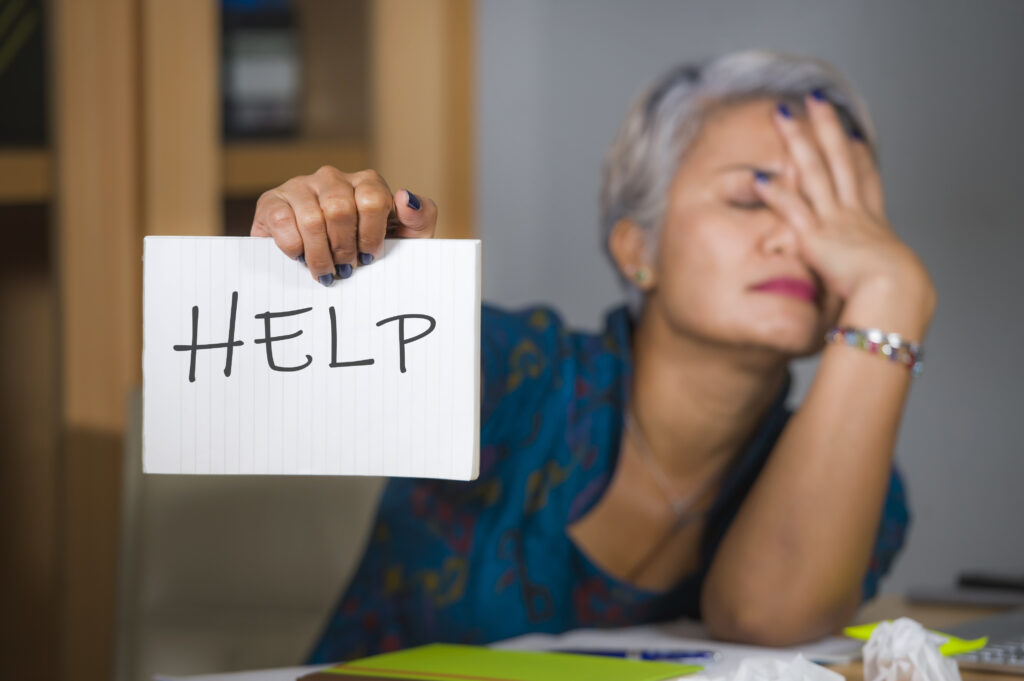 desperate and stressed attractive middle aged Asian woman holding notepad asking for help feeling overworked and exploited working at computer office desk suffering stress