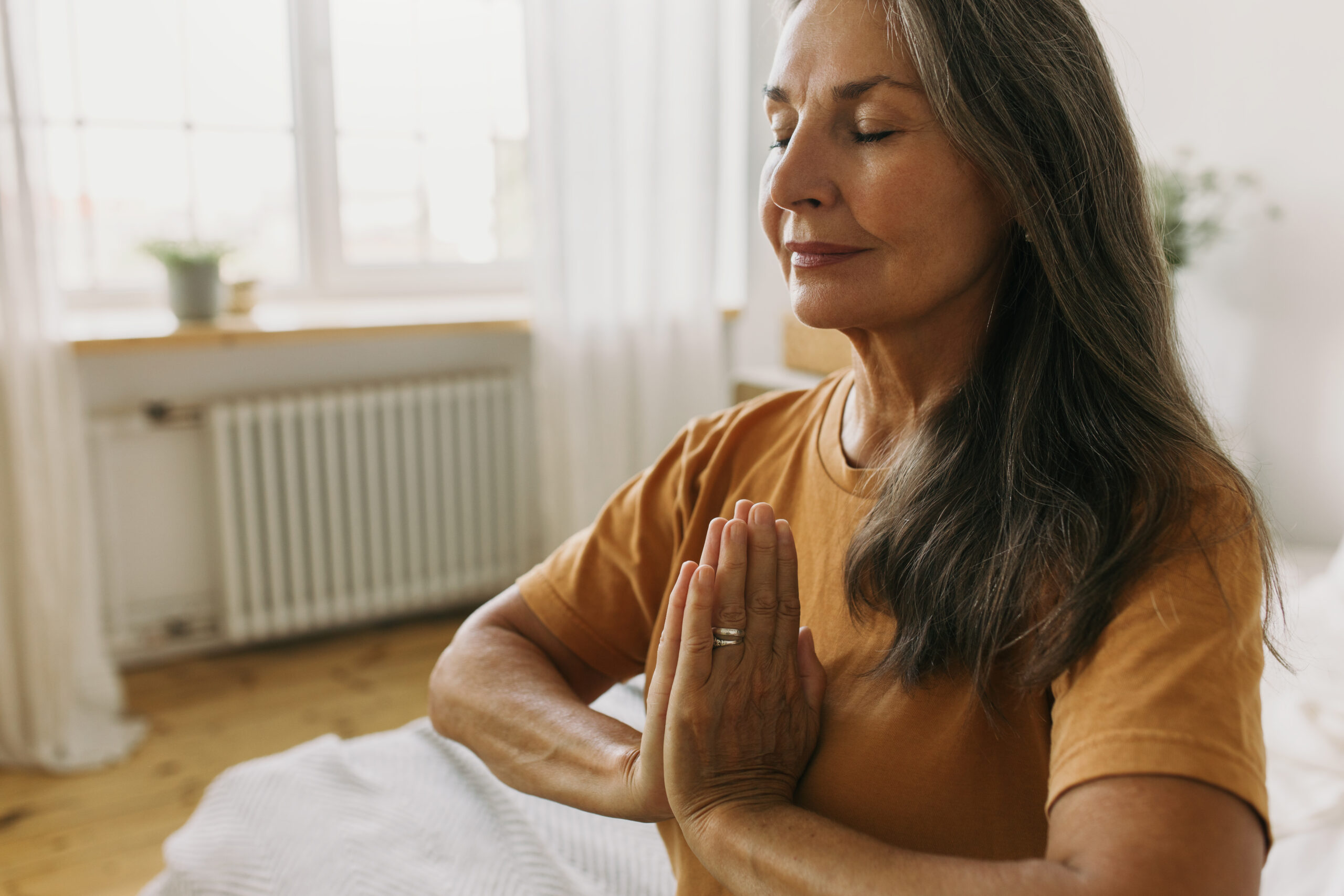 Calm middle-aged woman sitting in padmasana with eyes closed. Mature female holding hands pressed together in namaste and doing breathing exercises. Meditation and yoga idea